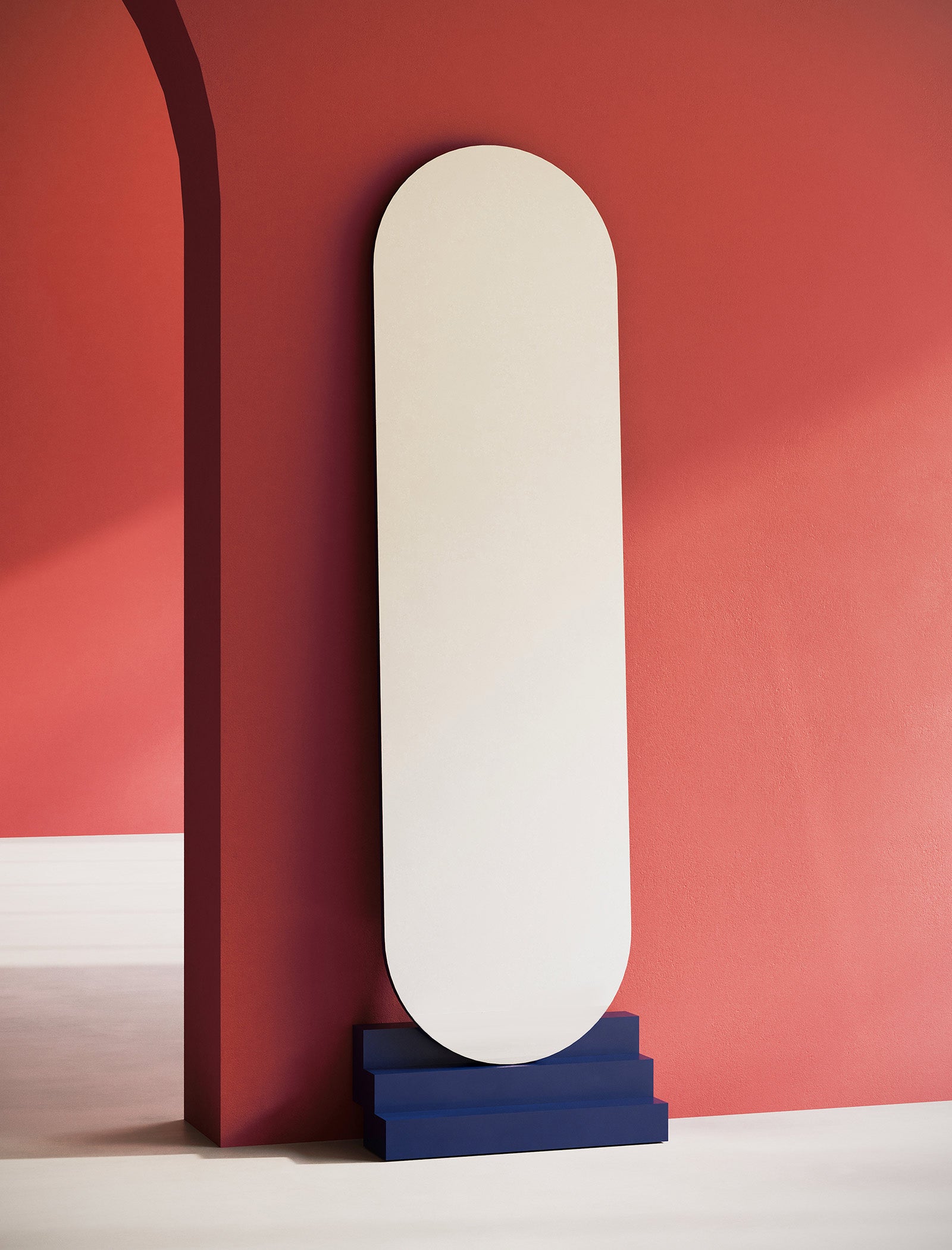 NONCHALANT Mirror L leaning securely against a red wall with a blue stepped base in minimalistic style.